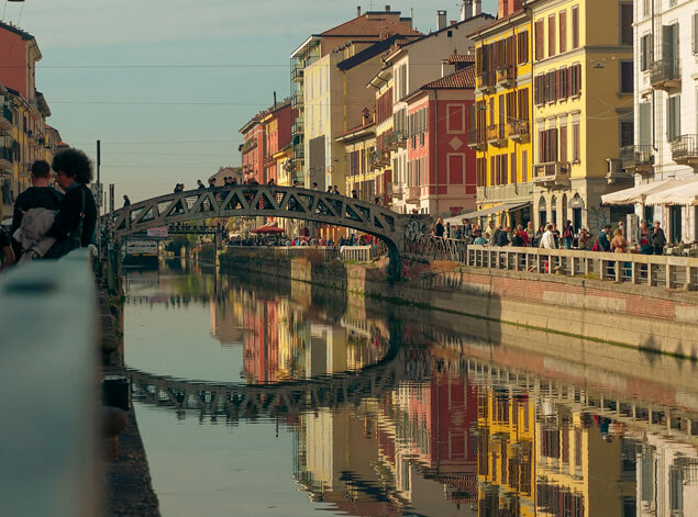 Milan: The Navigli canal at dusk, ochre facades and an iron footbridge mirrored in still water, figures on the bank