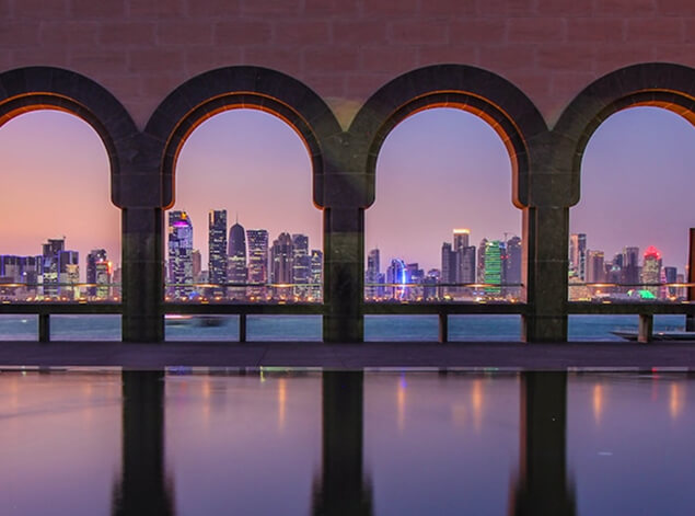 Doha: The West Bay skyline at dusk seen through three arches of the Museum of Islamic Art park, lights on the water