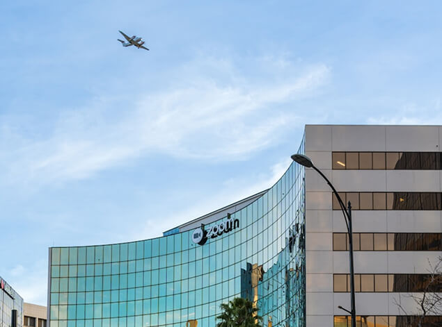 San José: A curved glass office building under broad cloud, a turboprop crossing the sky above it