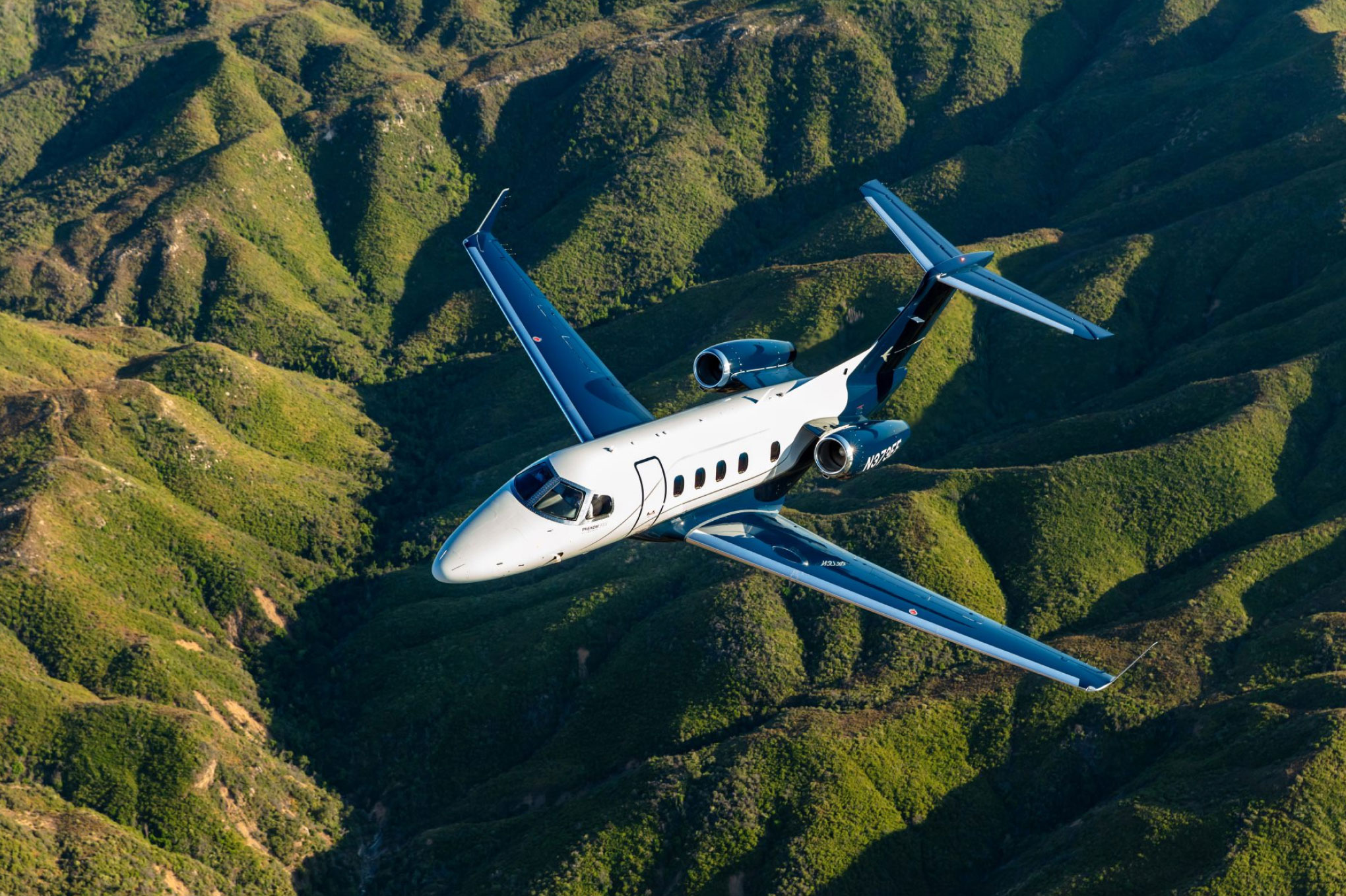 Embraer Phenom 300E in flight over green mountain ridges